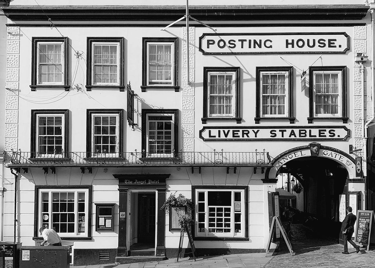 The Angel Hotel, Guildford. A black-and-white photograph of a historic two-story building with ornate window frames and wrought-iron balconies. Large signs read “POSTING HOUSE” and “LIVERY STABLES,” with an arched passageway labeled “ANGEL GATE” on the right. The entrance is marked “The Angel Hotel,” and pedestrians, signs, and street fixtures line the foreground.