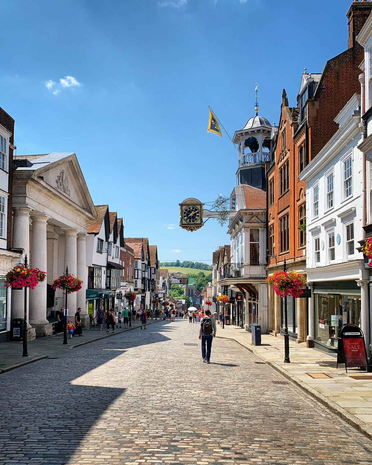 A distant view along Guildford's historic High Street with Tunsgate to the left, the Guildhall to the right and in the far distance the green hills and woods of The Mount. Vibrant hanging baskets line the pavement and a bright blue sky contrasts with the granite setts of the roadway. In the foreground, the profile of a man with a rucksack is walking away from the camera viewpoint.
